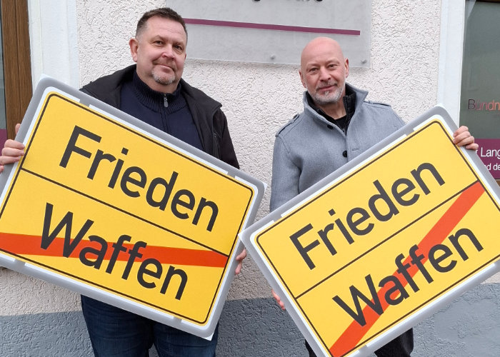 Das Bild zeigt zwei Männer die Schilder hochhalten. Auf diesen steht Frieden, Vernunft und Gerechtigkeit